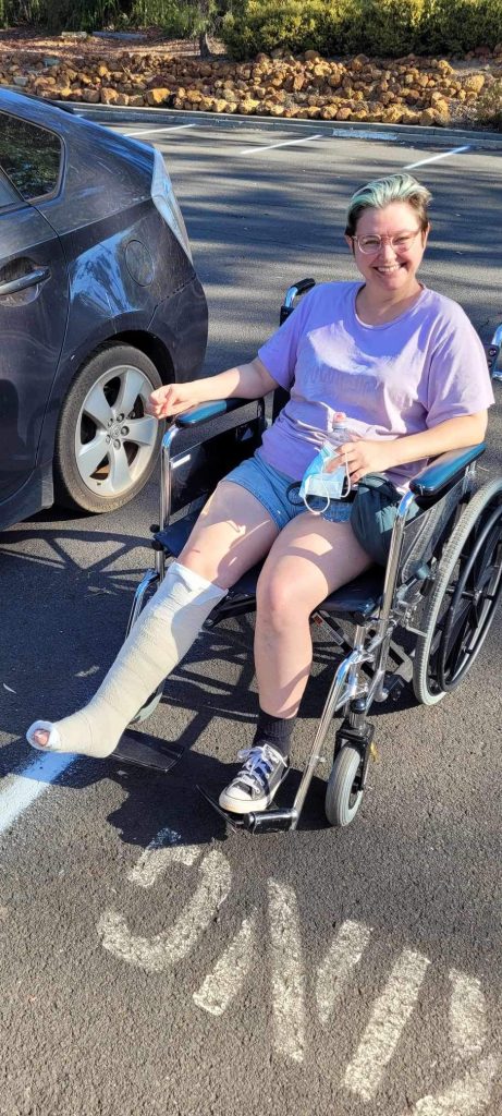 Me sitting in a wheelchair in a car park. I'm smiling, holding a face mask, and wearing a plaster cast on my right leg below the knee. 